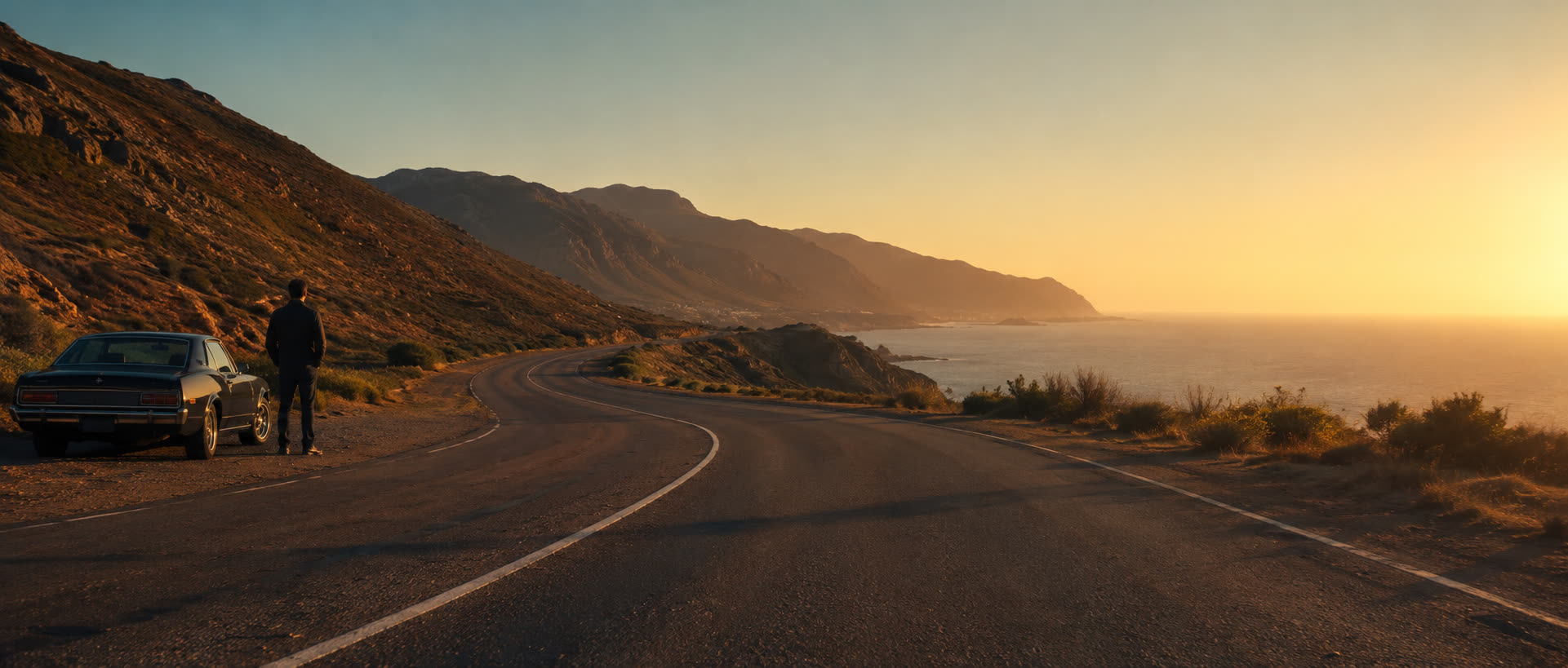 A traveller standing beside a parked car on an open coastal road at sunset, mountains and ocean stretching ahead